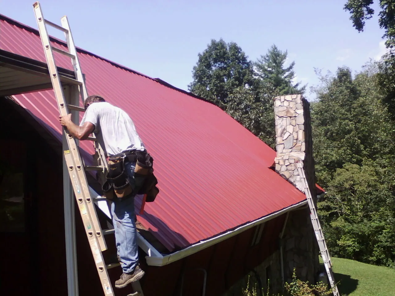 Roofer on a ladder beside a red metal roof and stone chimney — Soffit & Fascia Repair by Roofing Brothers in Clint